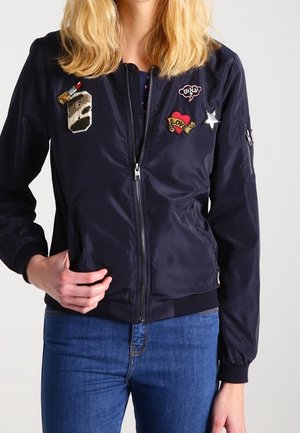 Vrouw draagt een marineblauwe bomberjack versierd met patches, waaronder een lipstick, hart met "LOVE", ster en spraakballon met "why?".