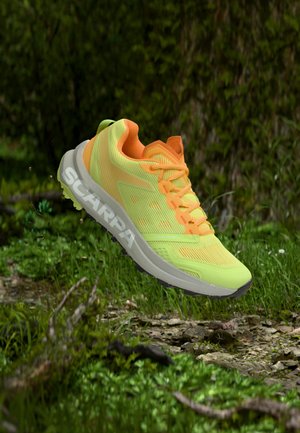 Lysegul og orange trail-løbesko i luften over en skovbund med græs og små sten, der viser SCARPA-logoet på sålen.