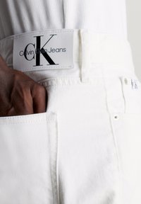 White denim jeans featuring a visible CK logo leather patch on the waistband, with a front pocket and a textured finish.