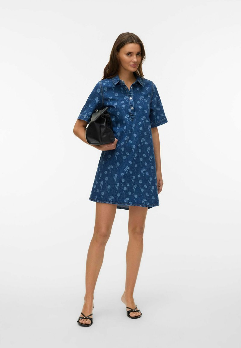 Femme debout en robe en jean à motif floral bleu, tenant un sac à main noir, portant des sandales à talons noires sur un fond blanc uni.