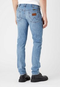 Pessoa vestindo jeans slim-fit azul claro com bolsos traseiros e sapatos pretos de atacador, em pé sobre um fundo branco simples.