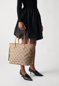 Beige taška přes rameno s hnědým vzorem, koženými detaily a dlouhými uchy, spárovaná s černou sukní a špičatými černými botami.