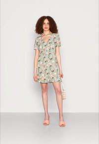 Vestido floral verde com mangas curtas, decote em V, bainha em camadas e um tecido leve. Combinado com sandálias de dedo laranja e uma pequena bolsa listrada.