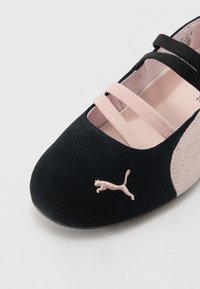 Black suede shoe with pink elastic straps and a light pink accent. Features a logo of a leaping cat in pink on the side. Smooth texture.