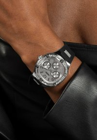 Strieborné hodinky s diamantovými akcentmi, textúrovaným šedým ciferníkom, tromi subciferníkmi a čiernym gumovým remienkom. Nosia sa na opálenej ruke, sčasti zakrytej látkou.