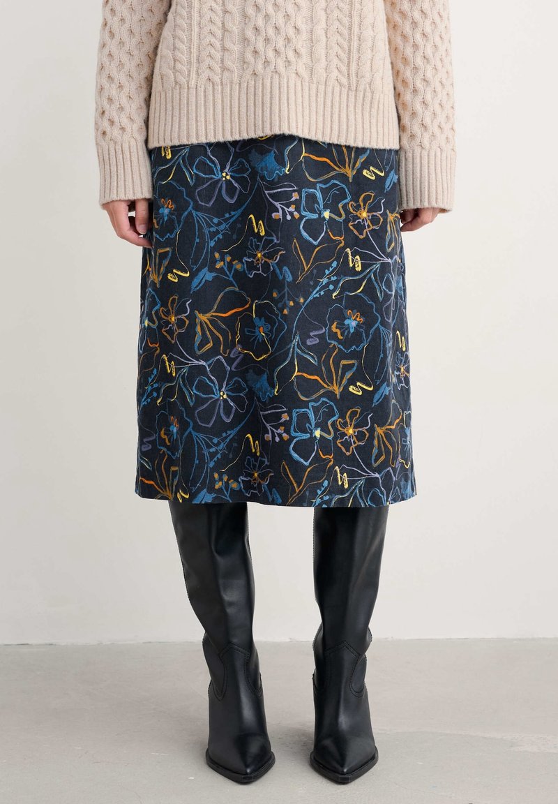 Jupe bleu marine longueur genou avec un motif floral coloré, associée à un pull beige en maille torsadée et des bottes en cuir noires montant jusqu'au genou.