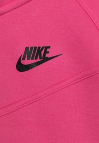 Růžová mikina se strukturovaným povrchem, s velkým černým logem Nike a zakřivenými švy. Látka je hladká a hustě pletená.