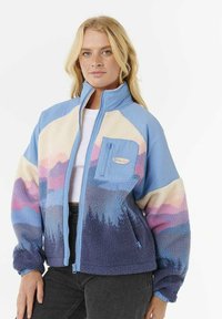 Kvinde med lyst hår iført en multicolor sherpa fleecejakke med blå, lilla, pink og creme nuancer, lynet halvvejs op, stående mod en hvid baggrund.