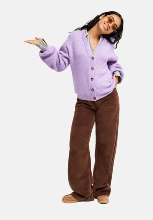 Femme aux longs cheveux foncés souriante, portant des lunettes de soleil, un cardigan violet, une chemise rayée, un pantalon en velours côtelé marron et des chaussures beige, debout avec une main levée.