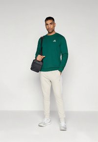 Sweatshirt verde, calças de treino com listra branca e sapatos cinza. Mala preta de ombro. Design simples com uma marcação subtil no peito.