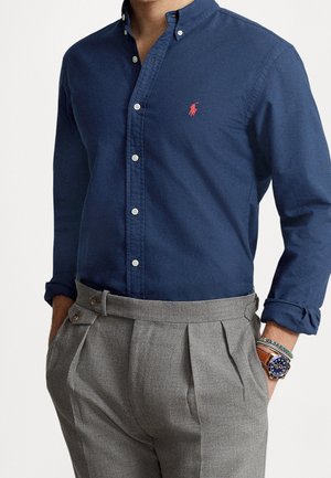 Mann trägt ein marineblaues Hemd mit rotem Logo und graue plissierte Hose, Hände in den Taschen und eine Armbanduhr mit Armbändern am linken Handgelenk.