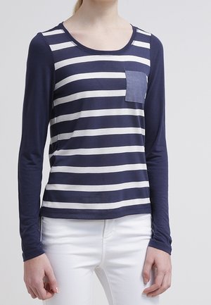 Vrouw die een navyblauw-wit gestreept shirt met lange mouwen draagt, voorzien van een effen navyblauw borstzakje, en witte broek.