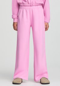 Pantaloni della tuta larghi di un rosa chiaro in morbido tessuto, con cintura elastica e tasche laterali. Indossati con scarpe slip-on beige.