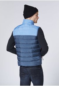 Padded vest in light blue and navy with horizontal stripes; features a stand collar and is worn over a black long-sleeve shirt.