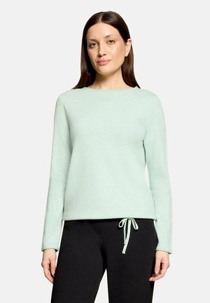 Femme aux cheveux lisses et foncés portant un pull léger vert menthe à manches longues et un pantalon noir avec un cordon noué.