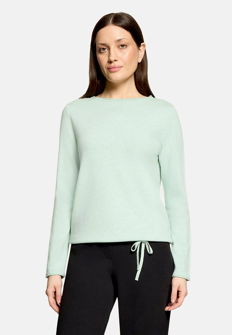 Femme aux cheveux lisses et foncés portant un pull léger vert menthe à manches longues et un pantalon noir avec un cordon noué.
