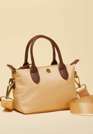 Kleine beige Handtasche mit dunkelbraunen Griffen und abnehmbarem beigem Schulterriemen vor hellem Hintergrund.