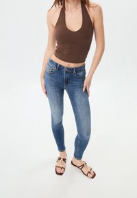 Mujer vestida con un top halter marrón, jeans skinny azules y sandalias marrones con acentos dorados, de pie contra un fondo blanco liso.