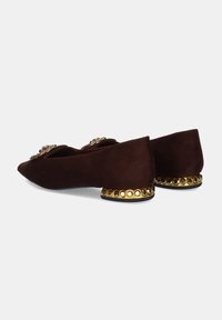 Zapatos planos de ante marrón con diseño de punta afilada, adornados con un tacón dorado con remaches y acentos decorativos en la parte superior.