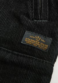 Zwart corduroy stof met een genaaid rechthoekig label met de tekst "Standard Issue Manufactured Goods ESTD 1982" in gele letters.