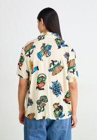Chemise à manches courtes en tissu crème, ornée de graphiques colorés et audacieux de masques, de bêtes et de motifs naturels, avec une coupe décontractée et un col.