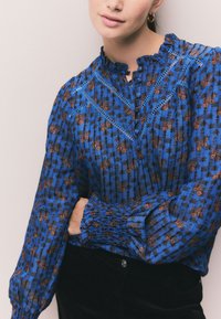 Niebieska bluzka z wzorem w kwiaty, z falbanianym dekoltem, zapinana na guziki, z marszczonymi rękawami, w połączeniu z ciemnym materiałem.