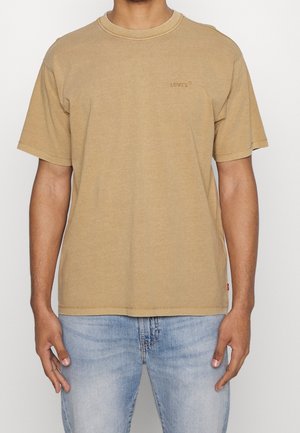Mężczyzna ubrany w prosty beżowy t-shirt Levi's z krótkim rękawem i jasnoniebieskie dżinsy, stoi na białym tle.
