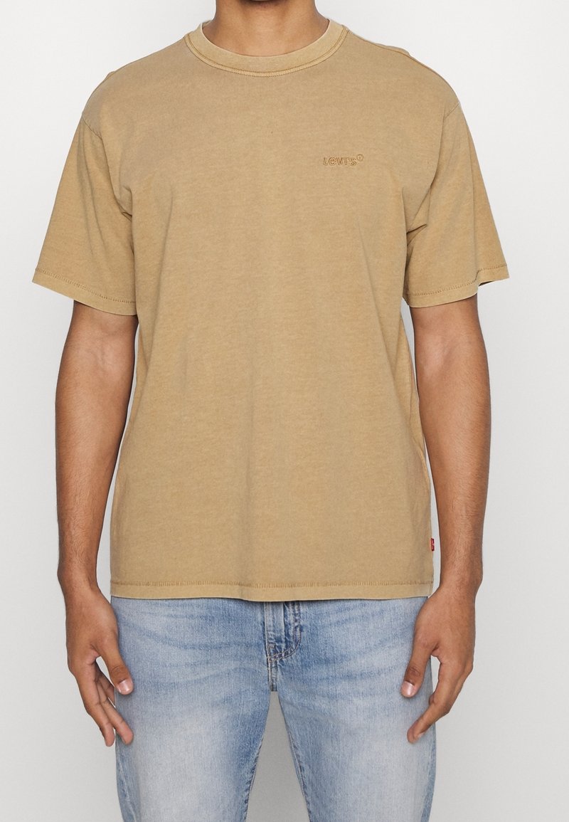 Homme portant un t-shirt Levi's beige uni à manches courtes et un jean bleu clair, debout devant un fond blanc.