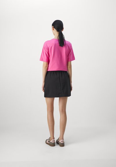 Kort rosa bomull t-skjorte med et croppet design, kombinert med et svart skjørt. Modellen har på seg mønstrede sko med tykk såle.