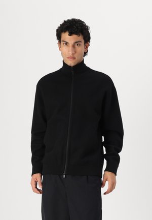 Schwarzer Reißverschluss-Pullover mit hohem Kragen und langen Ärmeln. Der Stoff wirkt weich, hat eine lockere Passform und gerippte Bündchen.