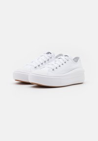 White canvas sneakers with a thick sole, lace-up front with metallic eyelets, rubber outsole, and textured detailing along the sides.