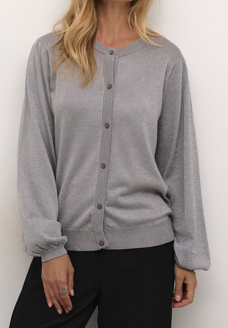 Femme portant un cardigan argenté scintillant à manches longues boutonné avec des manches bouffantes et un pantalon noir, debout devant un fond clair.