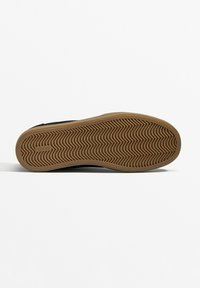 Black sneaker with a herringbone-pattern gum rubber sole, featuring a smooth upper and curved design. Logo label on the sole central area.