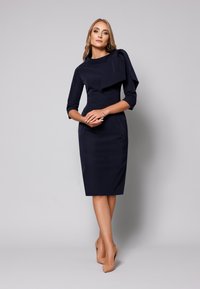 Granatowa dopasowana sukienka z rękawami o długości 3/4 i drapowanym akcentem na ramieniu, połączona z cielistymi szpilkami. Gładka struktura.
