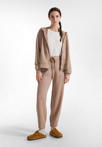 Coördinerende loungewear set in lichtbruin textiel. Inclusief rits-hoodie, broek met trekkoord, gecombineerd met bruine instap schoenen.