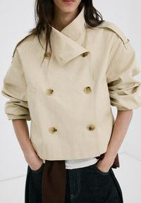 Femme portant une veste beige courte à double boutonnage avec des manches retroussées, mains dans les poches d'un pantalon en denim foncé, sur fond blanc.