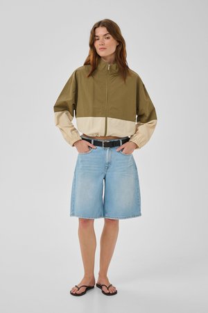 Naine kannab kahvatu oliivrohelist ja beeži kahetoonilist lukuga jopet, hele siniseid teksapükse, musta vööd ja musti plätusid, seisab käed taskutes.