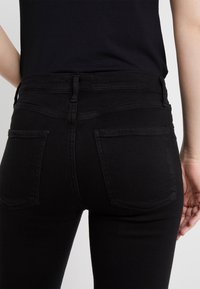 Henkilö, joka on pukeutunut tiukkoihin mustiin farkkuihin ja mustaan paitaan, näkyy vyötäröstä alas puoliväliin reisiä, takataskut ja vyölenkit ovat näkyvissä.