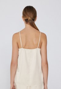 Débardeur beige clair avec de fines bretelles réglables, un dos légèrement froncé et des accents en dentelle le long de l'ourlet, fabriqué à partir d'un tissu lisse.