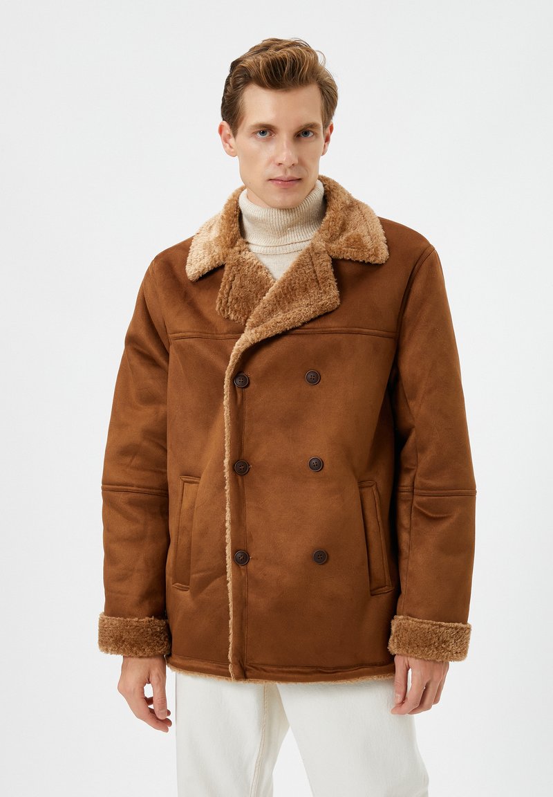 Mand iført en brun shearlingjakke med stor krave og manchetter over en beige turtleneck og hvide bukser, står mod en ensfarvet baggrund.