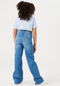Vaqueros de mezclilla azul acampanados con bolsillos traseros y una etiqueta de cuero marrón en la cintura, combinados con una camiseta de color azul claro.