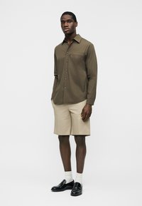 Mand iført olivengrøn langærmet skjorte med knapper, beige knælange shorts, hvide sokker og sorte loafers, stående mod en ensfarvet baggrund.