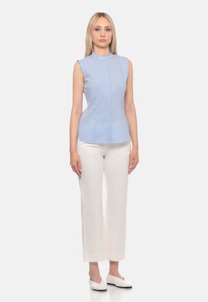 Donna in piedi che indossa una camicetta smanicata azzurro chiaro, pantaloni bianchi corti e ballerine bianche contro uno sfondo uniforme.