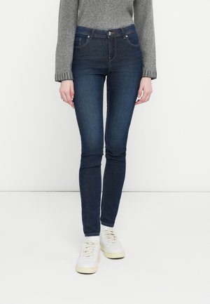 Jean skinny bleu foncé en denim, avec taille haute, deux poches avant et fermeture par bouton, associés à des baskets blanches.