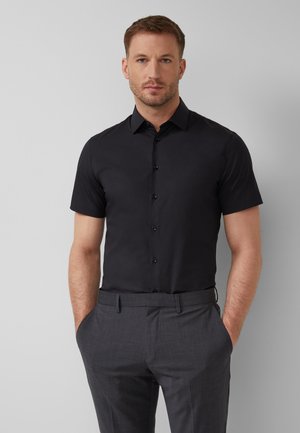 Mężczyzna o krótkich włosach, ubrany w czarną, krótką, zapinaną na guziki koszulę włożoną do szarych eleganckich spodni, stoi z rękami w kieszeniach.