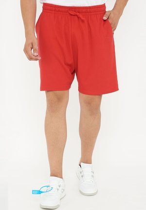 Mann trägt hellrote elastische Shorts mit Kordelzug und weiße Turnschuhe, steht vor weißem Hintergrund.