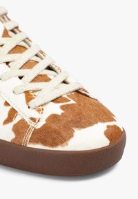 Zapatilla con parte superior de cuero de vaca que presenta un patrón marrón y blanco, cordones de un beige claro y una suela de goma con un acabado texturizado en marrón oscuro.