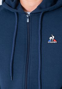 Felpa zip con cappuccio di colore navy realizzata in morbido tessuto, dotata di un cappuccio regolabile con cordino e un piccolo logo ricamato con accenti blu e rossi sul petto.