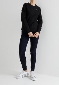 Felpa nera girocollo con polsini a costine, abbinata a jeans skinny blu scuro e sneaker bianche. Design semplice con dettaglio logo sul petto.