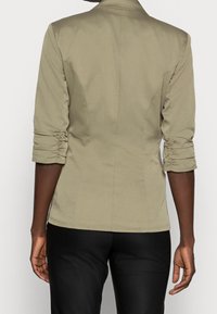 Blazer kaki clair avec manches trois-quarts froncées, coupe ajustée et texture lisse. Porté avec un pantalon noir, mettant en valeur une silhouette cintrée.